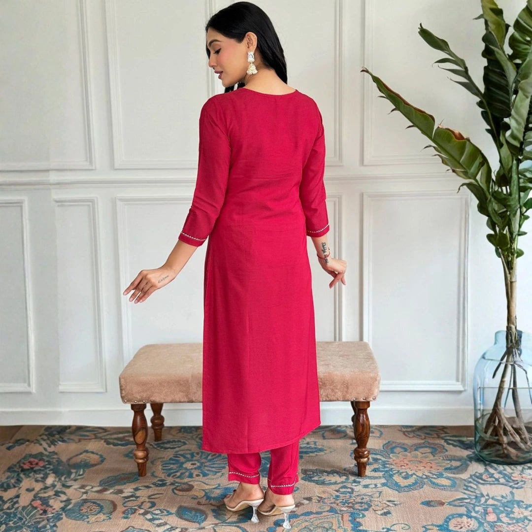 Woman in a red kurta standing in a room with a plant and bench.